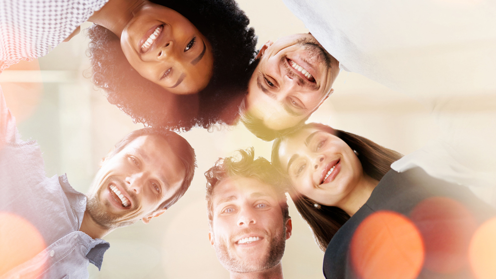 A group of men and women of various ethnicities huddled around in a circle looking down at the camera.