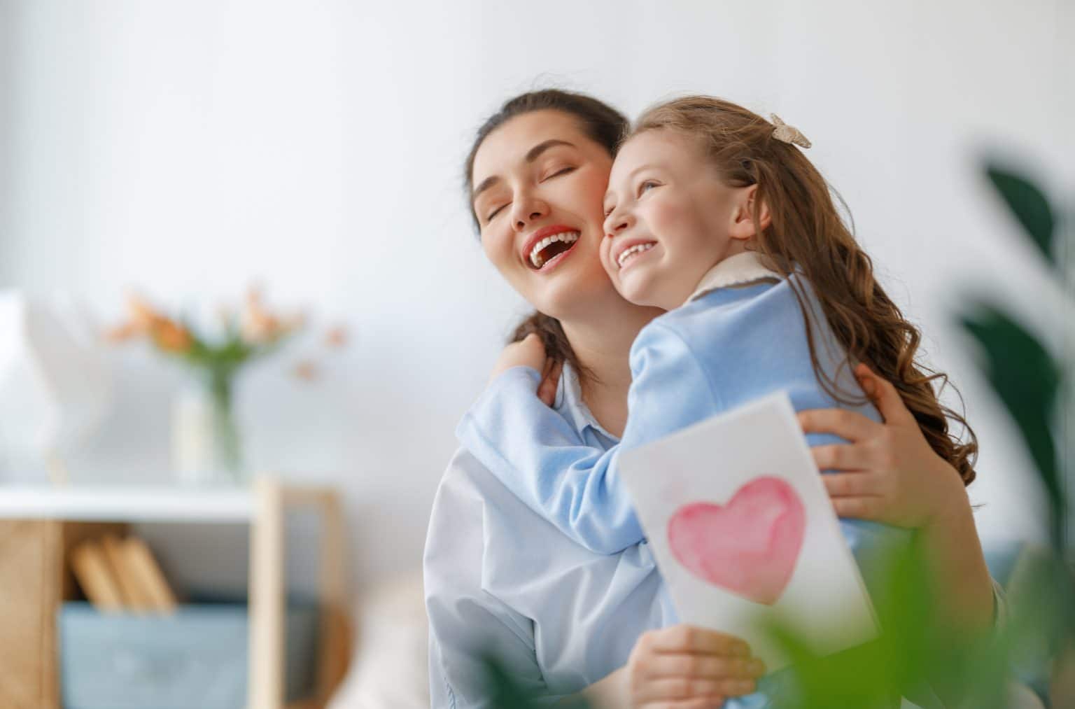 Happy mother's day. Child daughter is congratulating mom and giving her a homemade white card with a pink heart drawn on the front of it. Mum and girl smiling and hugging. Family holiday and togetherness.