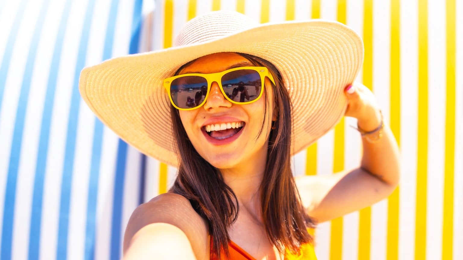 A happy brunette girl wearing a large brimmed sun hat and sunglasses taking a selfie at the beach.