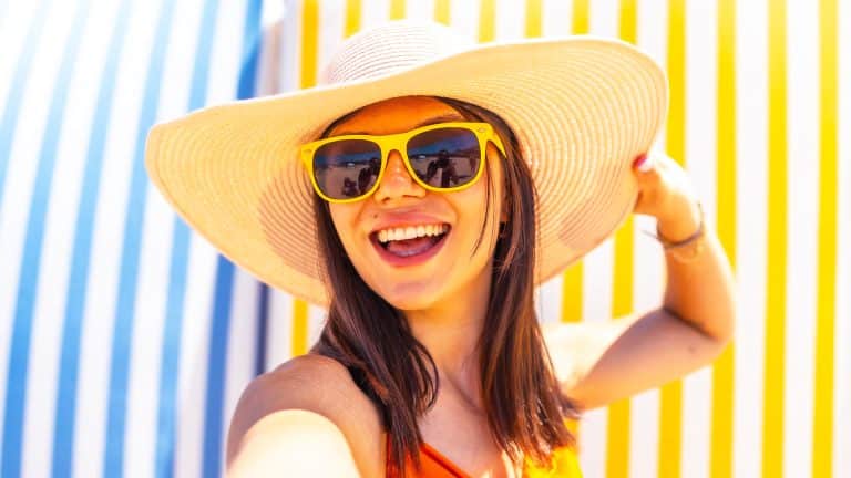 A happy brunette girl wearing a large brimmed sun hat and sunglasses taking a selfie at the beach.